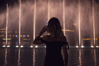 Rear view of woman standing against illuminated fountain in city