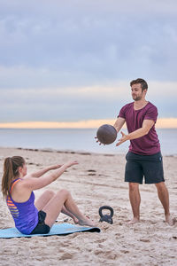 Trainer assisting woman exercising with medicine ball at beach