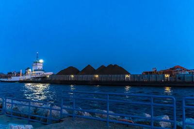 Illuminated buildings by sea against clear blue sky at night