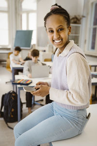 Portrait of happy schoolgirl holding smart phone while sitting at desk in classroom