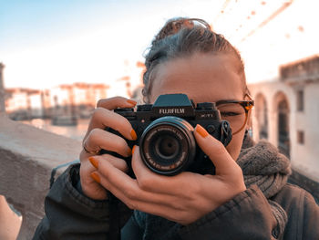 Close-up portrait of young woman holding camera against sky