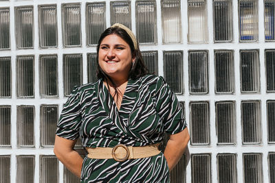 Young curvy female model in stylish black dress with diagonal striped print and belt standing against glass wall on street