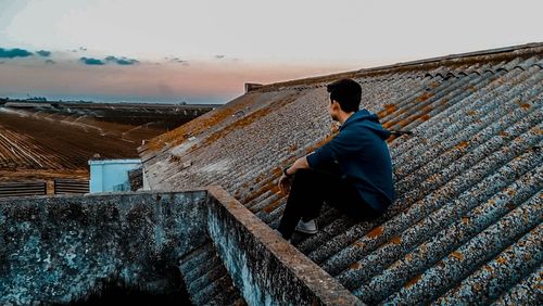 Rear view of man sitting on roof against sky