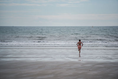Rear view of shirtless mature man running at beach against sky