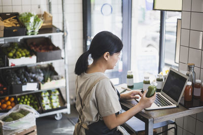 Asian female entrepreneur using laptop while holding juice bottle at store