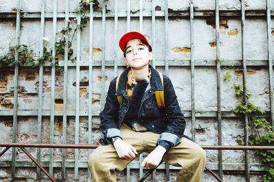 Portrait of boy wearing cap while sitting on railing against wall