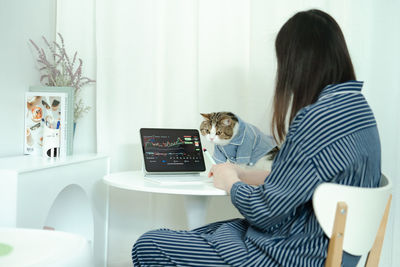 Woman trader in pajamas cloth work and check graph online from home and play with her cat