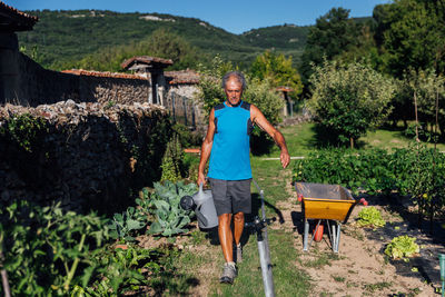 Full body front view of male farmer with watering can while standing near wall with plants