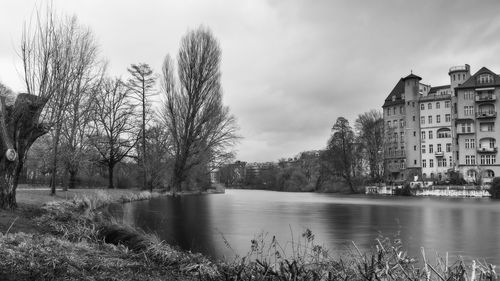 Bare trees by river against sky in city