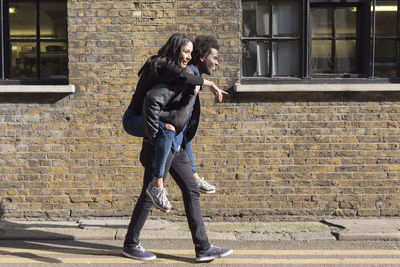 Young man carrying girlfriend piggyback at brick building