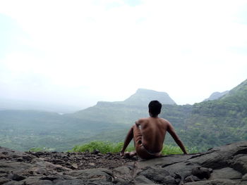 Rear view of shirtless man sitting on rock against mountains