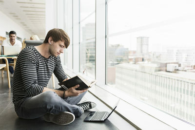 Male student reading book while sitting on floor at university library