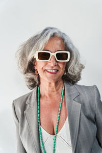 Positive elderly female in stylish jacket with beaded necklace and sunglasses smiling and looking at camera against white background