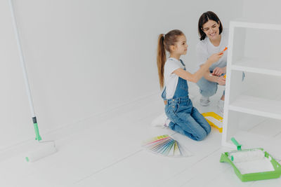 Full length of mother and daughter holding paint roller sitting at home