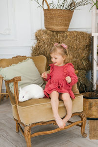 Three-year-old girl in a dress barefoot sits on a chair with a white rabbit in the living room
