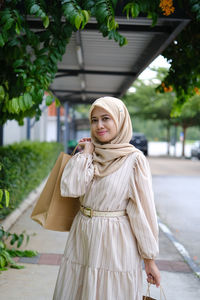 Young woman standing against shopping mall after shopping while holding paper bag 