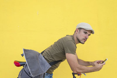 Man using phone while standing with bicycle against yellow wall