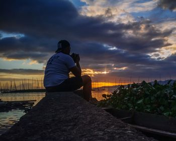 Man sitting on rock against sky during sunset