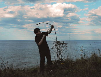 Boy taking aim with bow by sea against cloudy sky