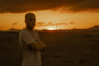 Portrait of young man standing at beach against sky during sunset