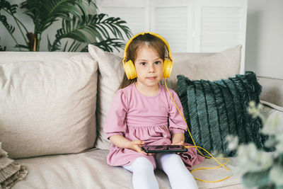 A little girl in yellow headphones sits on a sofa with a phone.