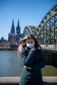 Portrait of young woman wearing mask standing against bridge over river