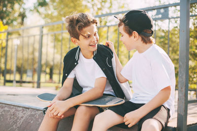 Two cute teenagers sit in a skatepark, relax after skateboarding and chat.