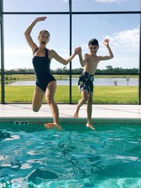 Full length of mother with son jumping in swimming pool against sky