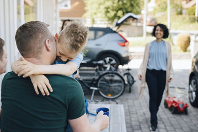 Rear view of father embracing son while woman walking towards house during weekend