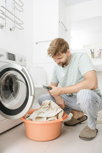 Man using smart phone by washing machine in bathroom at home