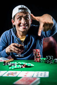 Young man pointing at poker table while holding whiskey against black background