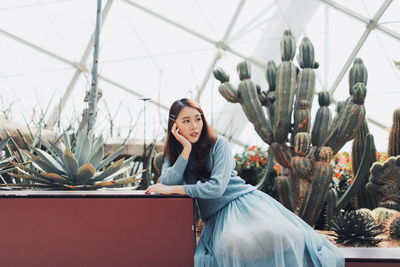 Thoughtful young woman sitting against plants in greenhouse