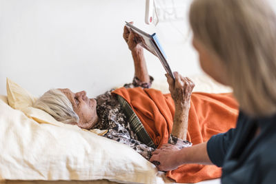 Woman taking care of old woman lying in bed reading book