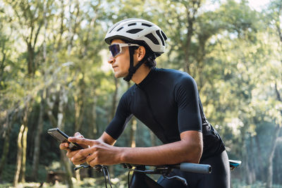 Male biker in helmet and cycling glasses text messaging on cellphone while sitting on bicycle in woods on sunny day