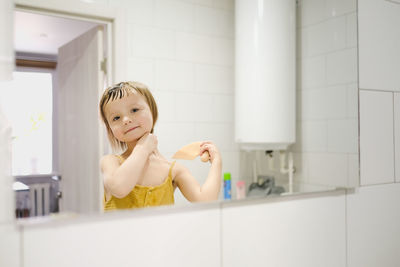 Child in yellow jumpsuit combs his own in front of mirror in bathroom