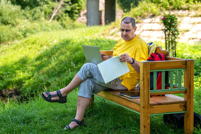 Mature man with laptop and documents working outside in garden, green home office concept.