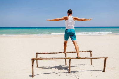 Man with arms outstretched balancing on wooden structure at beach during sunny day