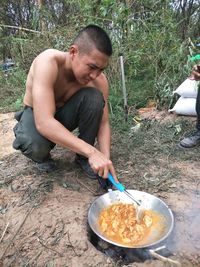 Shirtless man preparing meat on fire pit