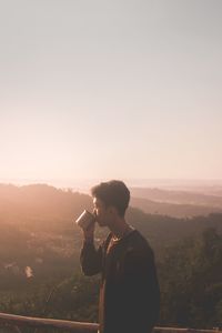 Side view of teenage boy drinking coffee on mountain peak against clear sky