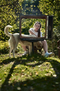 Young woman sitting with dog on grass seen through television set outdoors