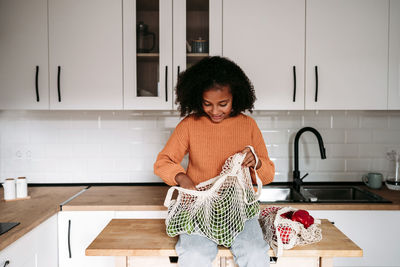 Smiling girl sitting with mesh bag of vegetables at home