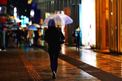 Full length rear view of woman with umbrella in illuminated city at night during monsoon
