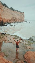 Rear view of shirtless mid adult man with arms raised standing at beach against sky