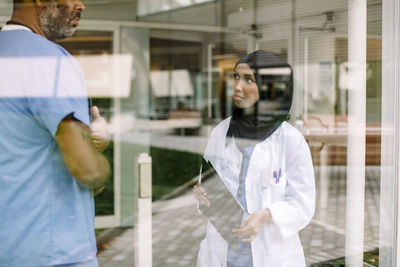 Young female nurse talking to colleague in hospital seen through glass window