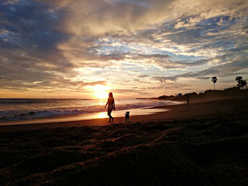 Silhouette woman with dog at beach against sky during sunset