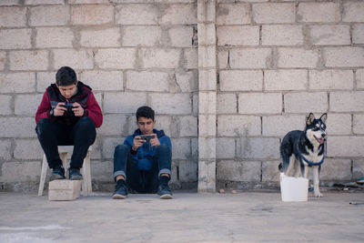 Friends using mobile phone by dog against brick wall