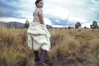 Beautiful woman standing with fur jacket on grassy field against cloudy sky