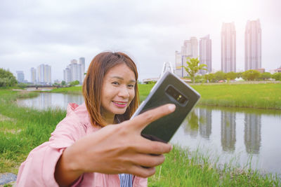 Smiling young woman using phone by lake against sky