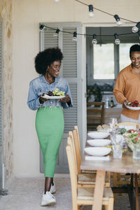 Happy woman with afro hairstyle walking with plate by dining table in backyard patio