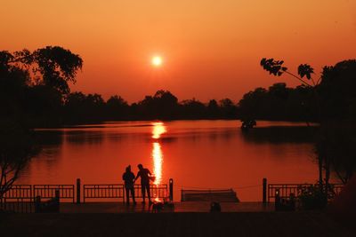 Silhouette people by lake against orange sky